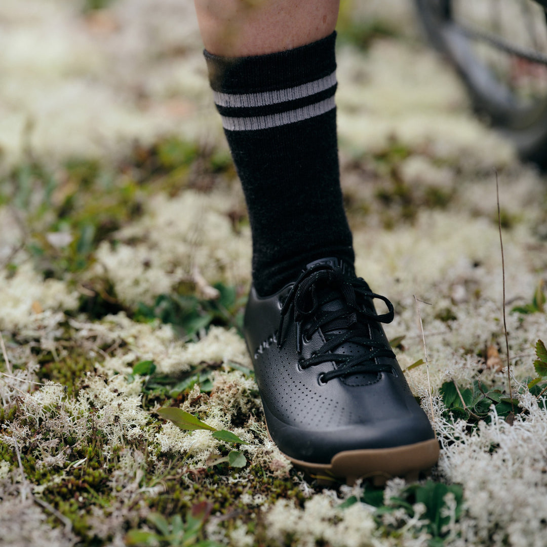 Pinebury Mountain Merino Wool Sock in Charcoal, Person wearing a black cycling shoe standing in white brush