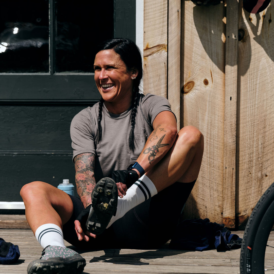 Pinebury Portland Short Sleeve Merino Wool Performance Tee in Granite, Woman smiling while sitting on ground in front of shed putting cycling shoes on before a ride