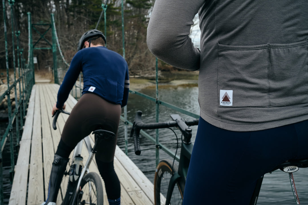 Pinebury Rangeley Long Sleeve Merino Wool Cycling Jersey in Granite, Close up of the pocket details on back of a cyclist stopped on a wood bridge over water. Other male cyclist in the background getting on his bike in a blue jersey and brown bibs
