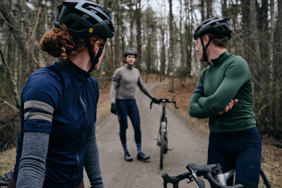Pinebury Rangeley Long Sleeve Merino Wool Cycling Jersey in Pine. Three cyclists stopped on a gravel road. A female cyclist red hair and a Male cyclist in a green jersey are look back, away from the camera, at another cyclist standing in the background next to his bike wearing a grey jersey.