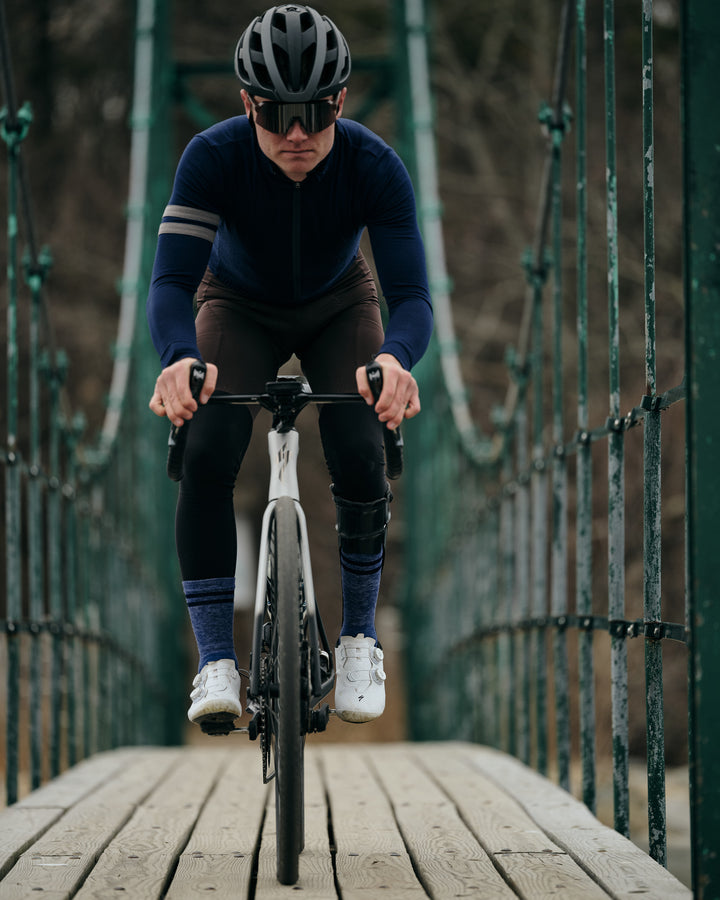 Pinebury Rangeley Long Sleeve Merino Wool Cycling Jersey in Atlantic Blue, Male cyclist riding straight towards the camera and looking focused over a suspended wooden bridge. He is wearing a blue jersey with brown bibs and white shoes with blue socks