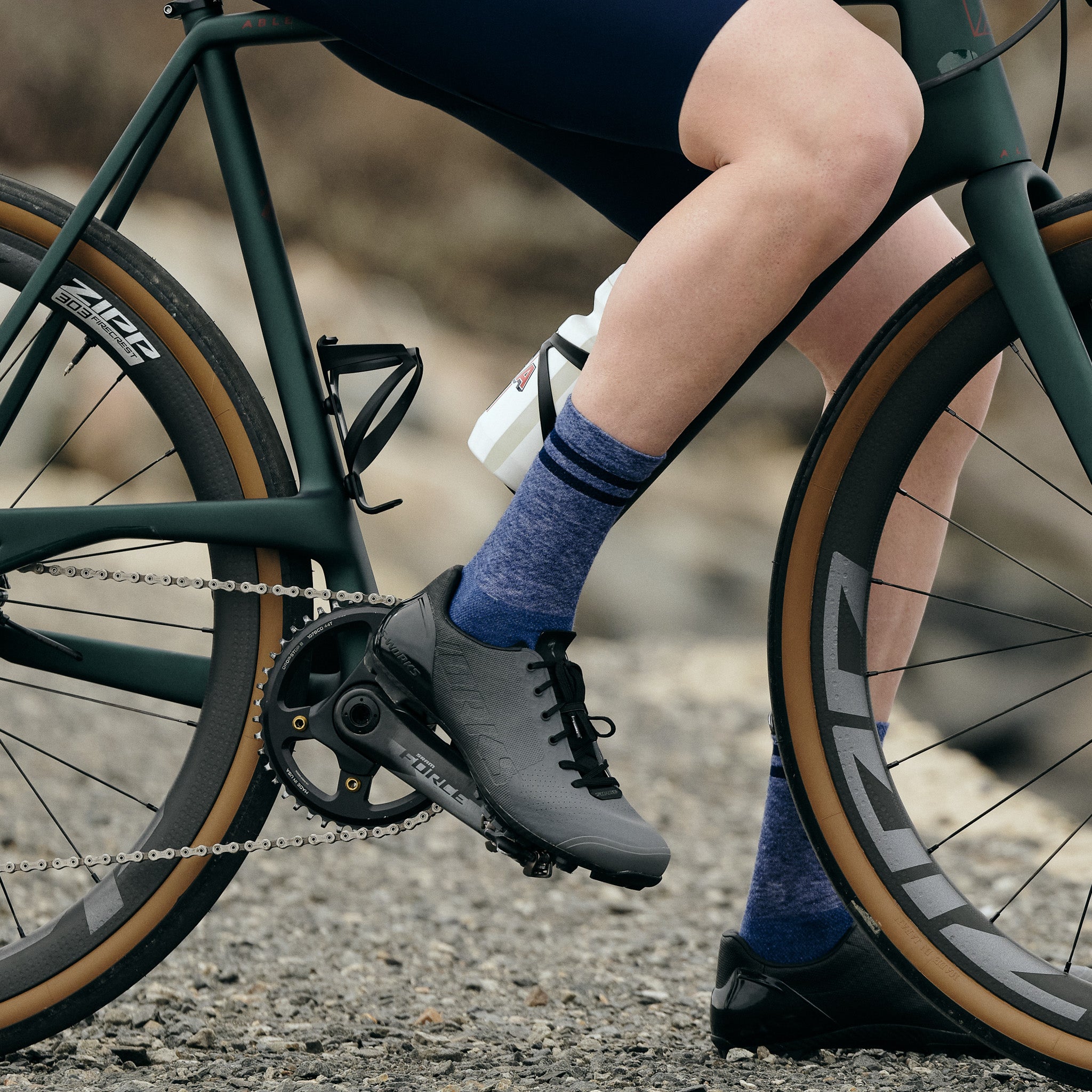 Pinebury Signature Merino Wool Sock in Blue. Close up side view of cyclist on a green gravel bike with their right foot on the pedal wearing a black and grey lace up cycling shoe and blue socks with two stripes at the top.