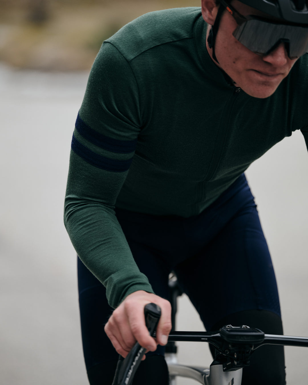 Pinebury Rangeley Long Sleeve Merino Wool Cycling Jersey in Pine. Close up of Male cyclist riding towards the camera out of the saddle. He is wearing a green cycling jersey with two blue stripes on the right bicep.