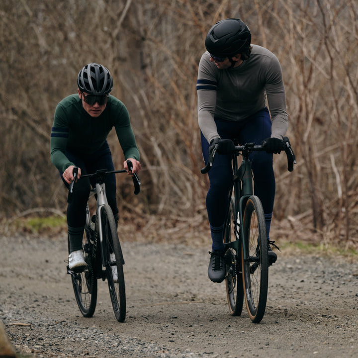 Pinebury Rangeley Long Sleeve Merino Wool Cycling Jersey in Granite, Two male cyclists riding towards the camera on a gravel road. One is wearing a grey long sleeve jersey and blue bottoms, the other in a matching green jersey.