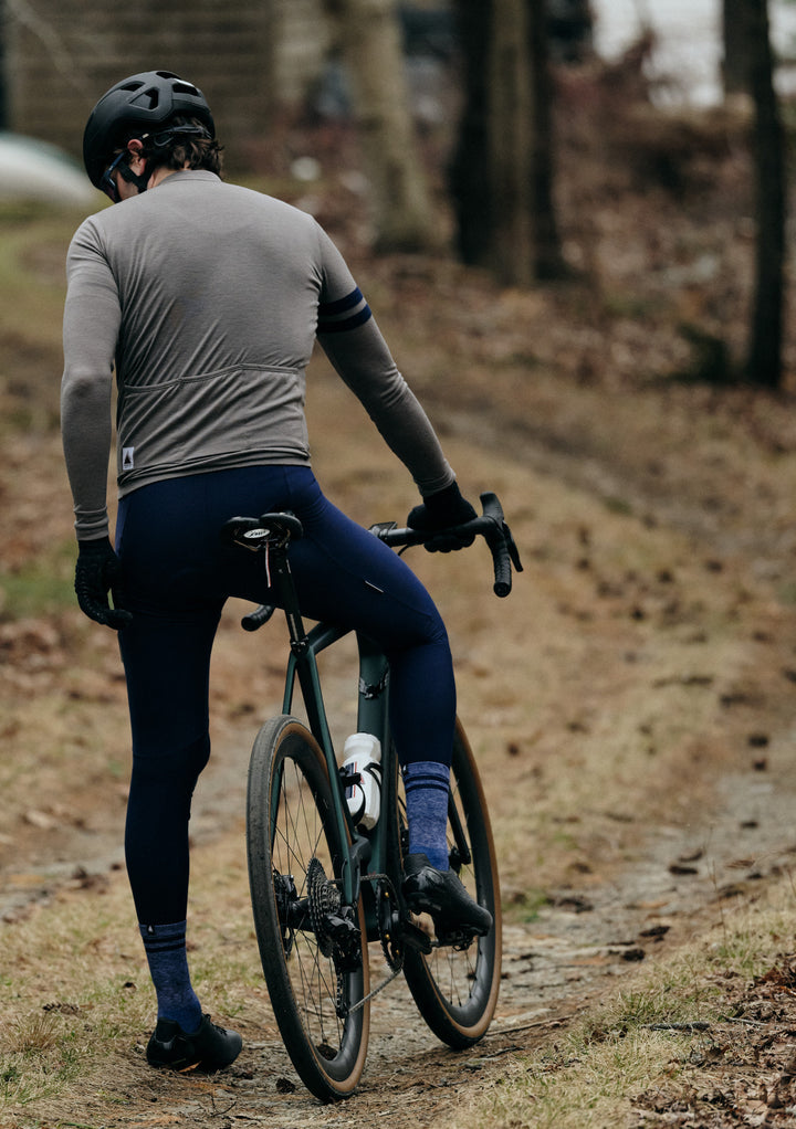 Pinebury Rangeley Long Sleeve Merino Wool Cycling Jersey in Granite. Male cyclist stopped on a gravel road in the fall. He is wearing a grey long sleeve jersey with blue bib tights and blue socks, riding a green gravel bike.