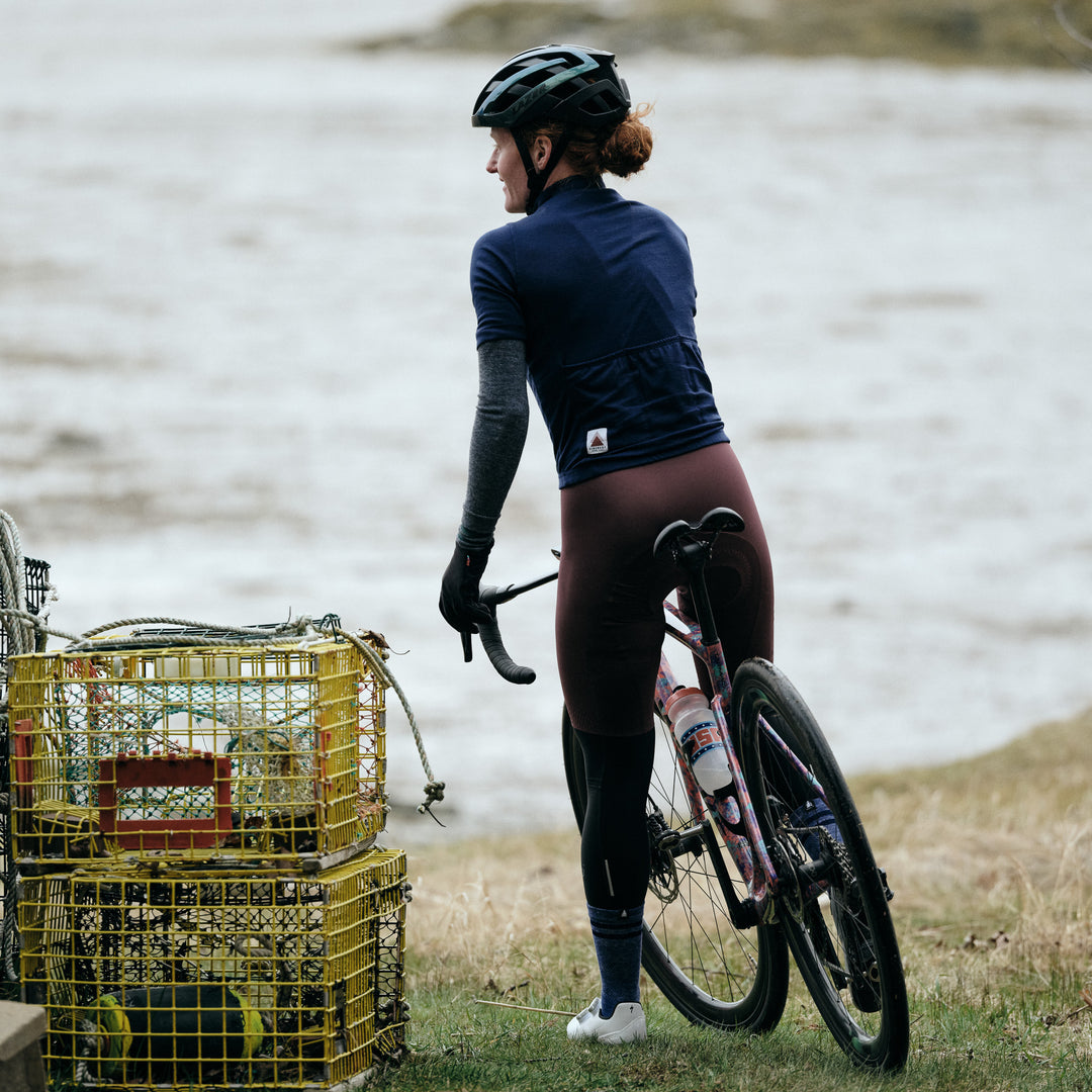 Pinebury Rangeley Short Sleeve Cycling Jersey in Atlantic Blue. Female cyclist stopped and looking into the water in the distance next to yellow lobster crates. She has red hair and is wearing a blue short sleeve jersey and brown bibs.
