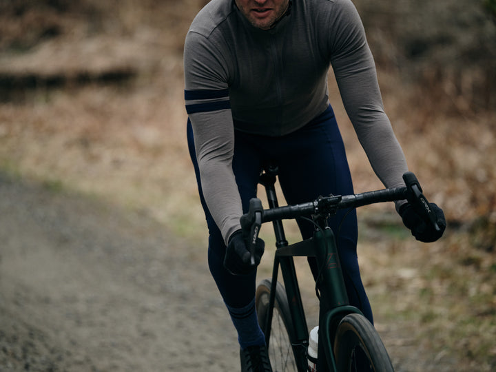 Pinebury Rangeley Long Sleeve Merino Wool Cycling Jersey in Granite. Close up of male cyclist riding towards the camera on a gravel road. He is turning slightly in the drops while riding a green gravel bike, wearing a grey jersey and blue bib tights.