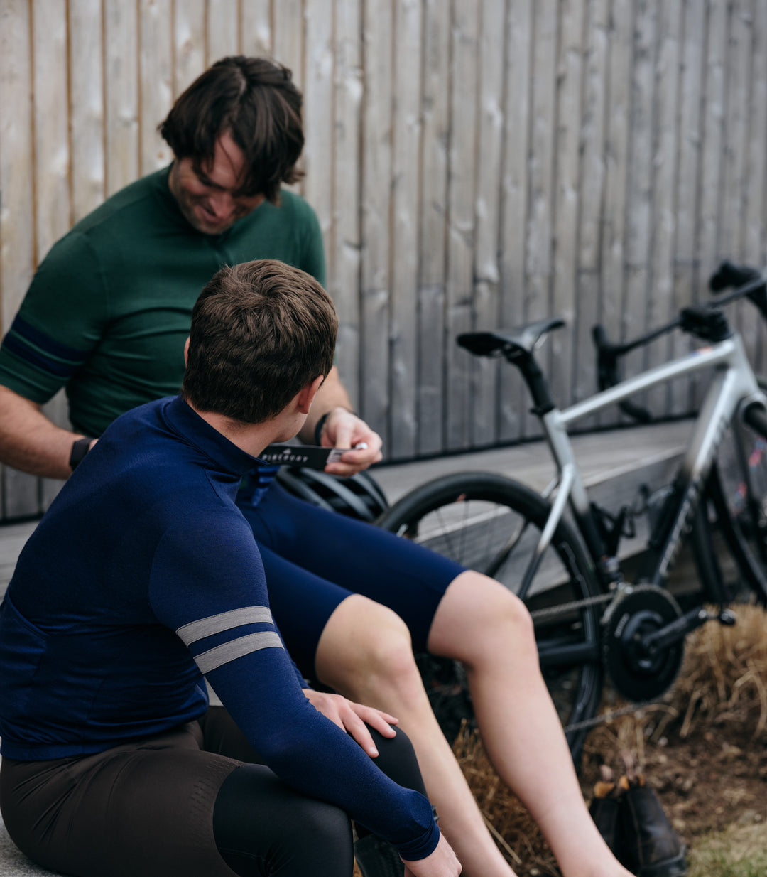 Pinebury Rangeley Long Sleeve Merino Wool Cycling Jersey in Atlantic Blue, Two male cyclist sitting and smiling in conversation while they get ready. A black and silver bike is leaning against the shed in the background.