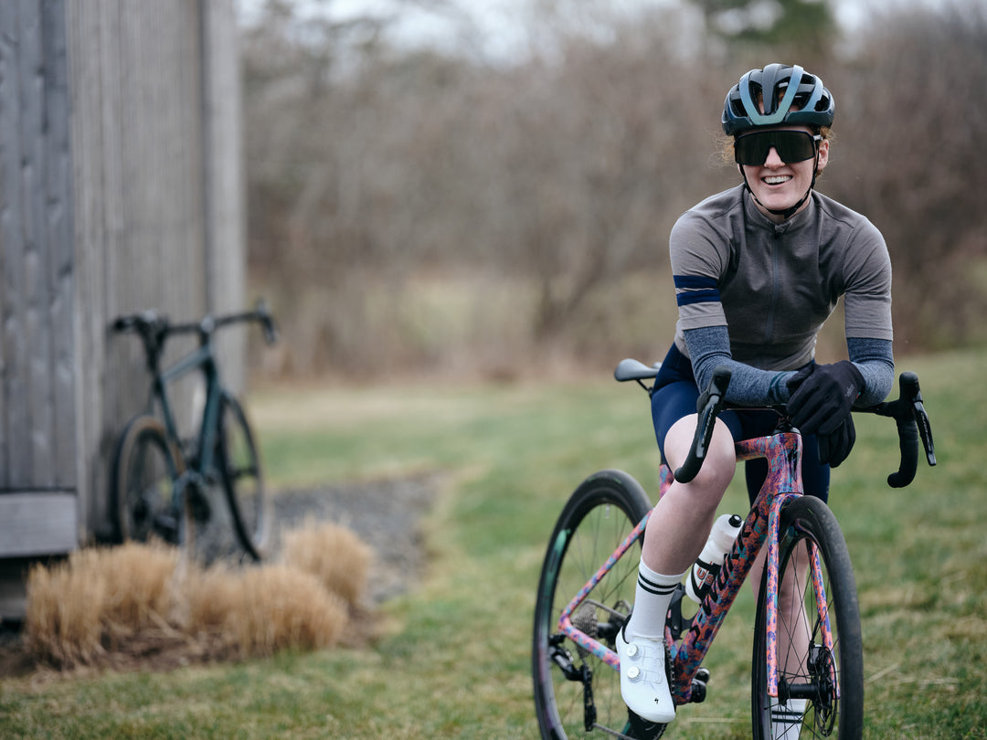 Pinebury Rangeley Long Sleeve Merino Wool Cycling Jersey in Granite. Female cyclist smiling at the camera while sitting on the top tube of her colorful pink bike. Wearing a grey short sleeve jersey and grey arm warmers, white shoes and socks.