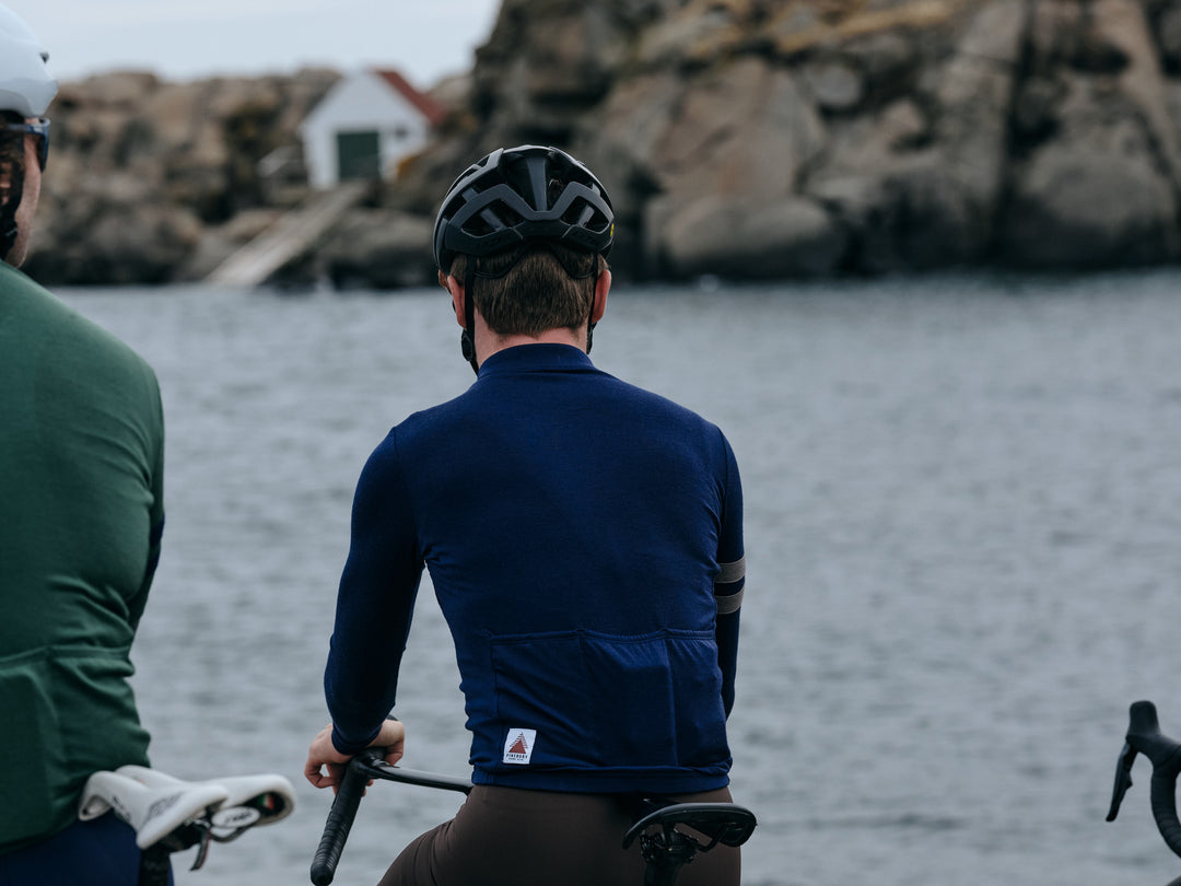 Pinebury Rangeley Long Sleeve Merino Wool Cycling Jersey in Atlantic Blue, Back view of a male cyclest looking towards an out of focus rocky coast. Wearing a blue long sleeve jersey with brown bibs and a black helmet.  