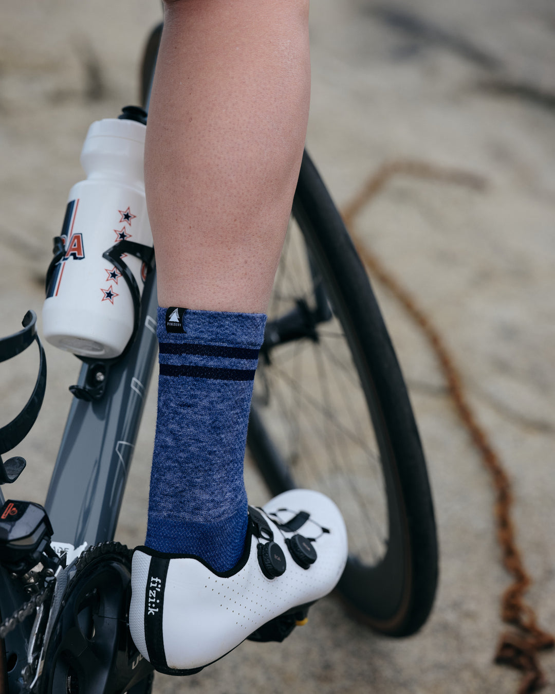 Pinebury Signature Merino Wool Sock in Blue. Close up rear shot of cyclist riding with right foot on the pedal in white shoes with blue socks.