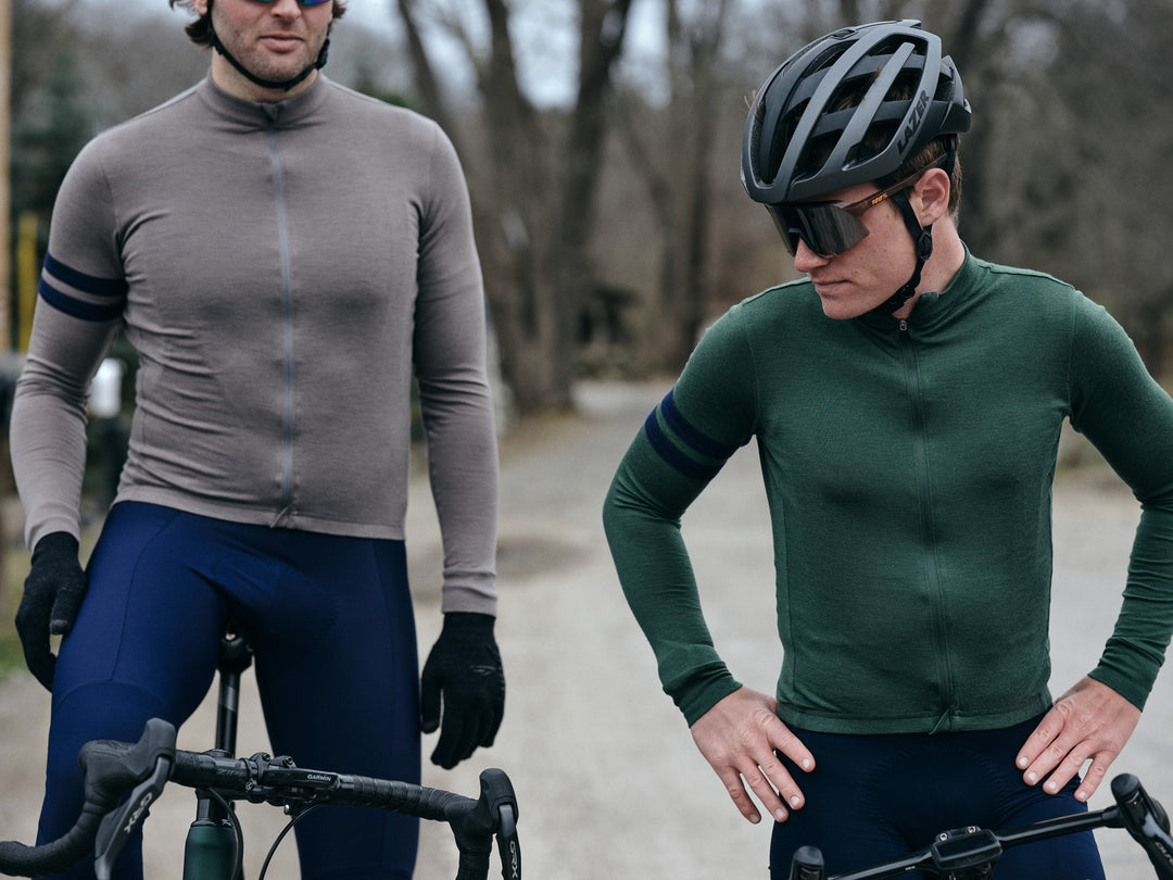 Pinebury Rangeley Long Sleeve Merino Wool Cycling Jersey in Granite, Two male cyclist standing over their bikes pensively. One rider is wearing a grey long sleeve jersey with blue bibs and the other has a green long sleeve jersey with a black helmet