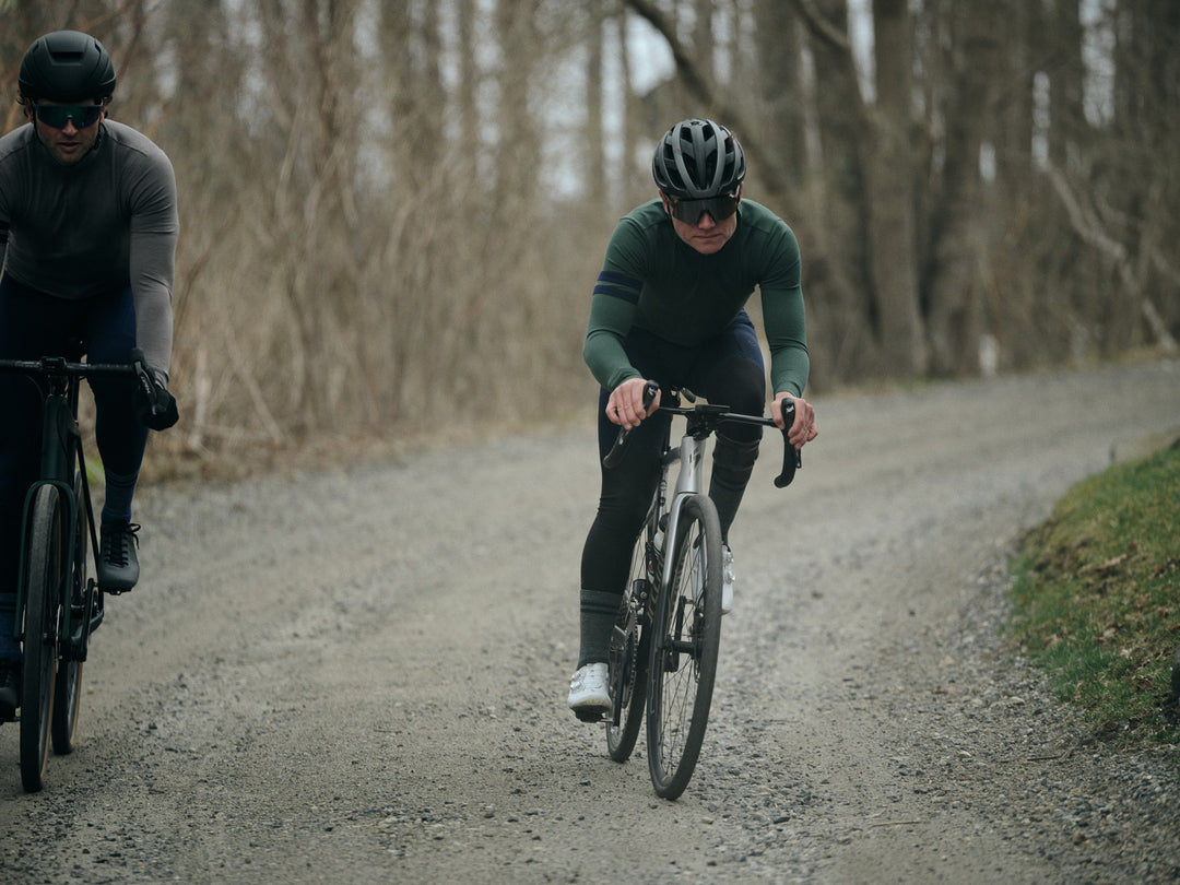 Pinebury Rangeley Long Sleeve Merino Wool Cycling Jersey in Pine. Two male cyclists riding down a gravel road with a slight decline towards the camera. The main cyclist in frame is wearing a green long sleeve jersey with blue stripes and green socks with white shoes.