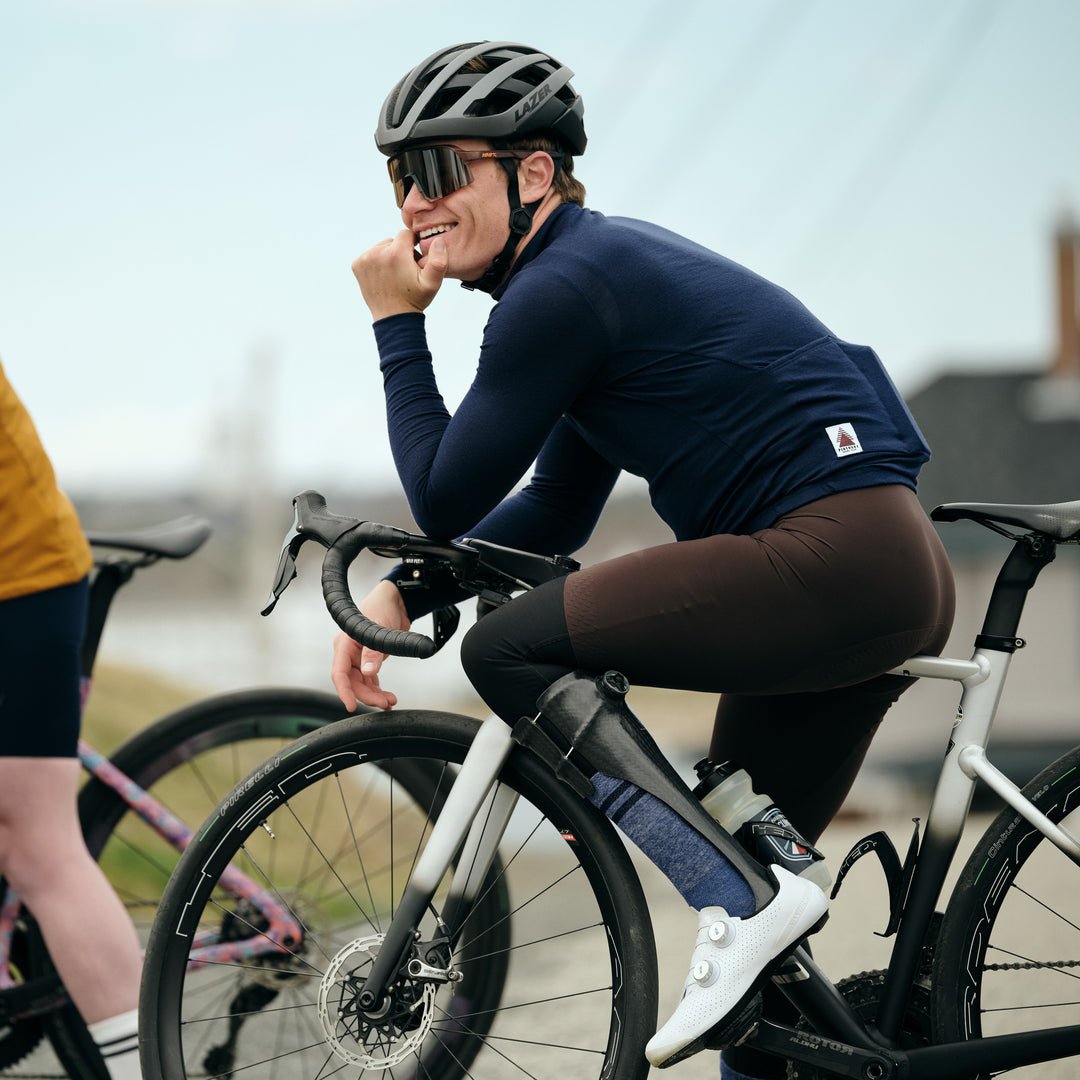 Pinebury Rangeley Long Sleeve Merino Wool Cycling Jersey in Atlantic Blue, Male cyclist stopped, sitting on the top tube of his bike smiling and looking to the distance, has brown bib shorts on and a carbon fiber lower leg brace with a blue sock showing through and white shoes