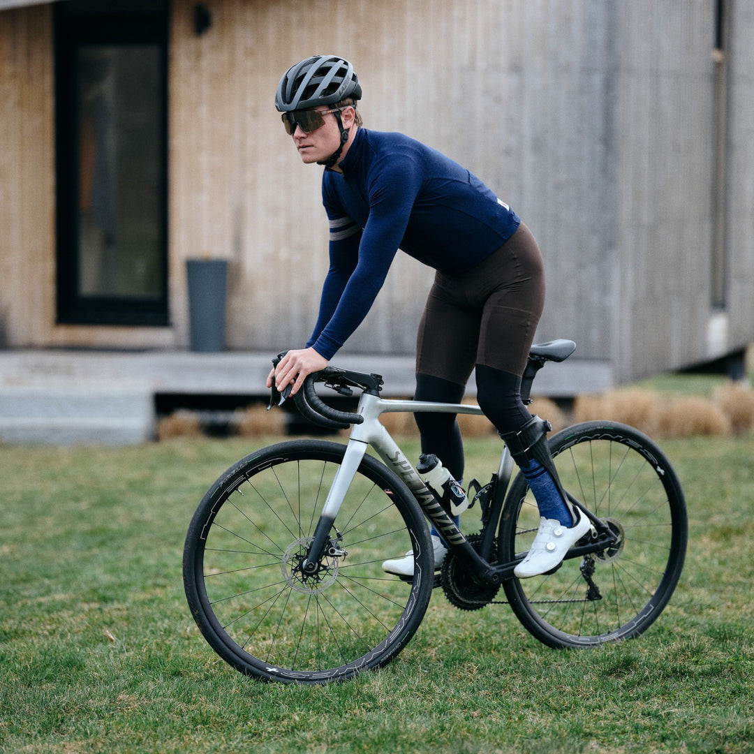 Pinebury Rangeley Long Sleeve Merino Wool Cycling Jersey in Atlantic Blue, Male cyclist doing a track stand in a field in front of a shed. He is wearing a blue long sleeve jersey with brown bibs, blue socks, and white shoes on a specialized road bike